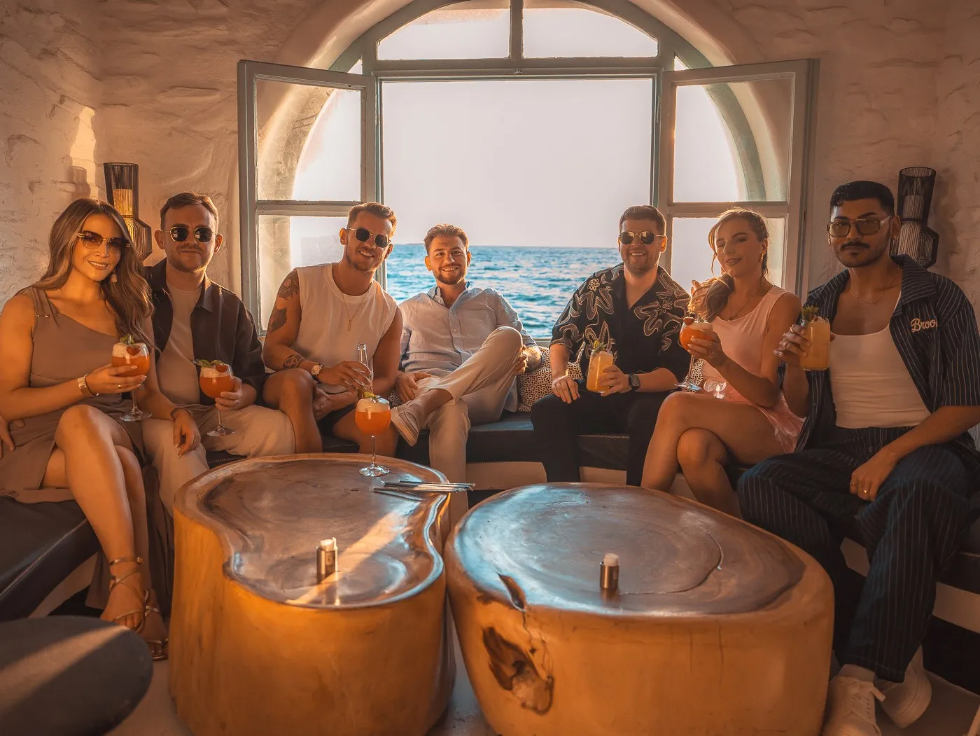 Eight guests seated around the Rotonda Table at BAO'S with cocktails, the arched window open to the sea behind them