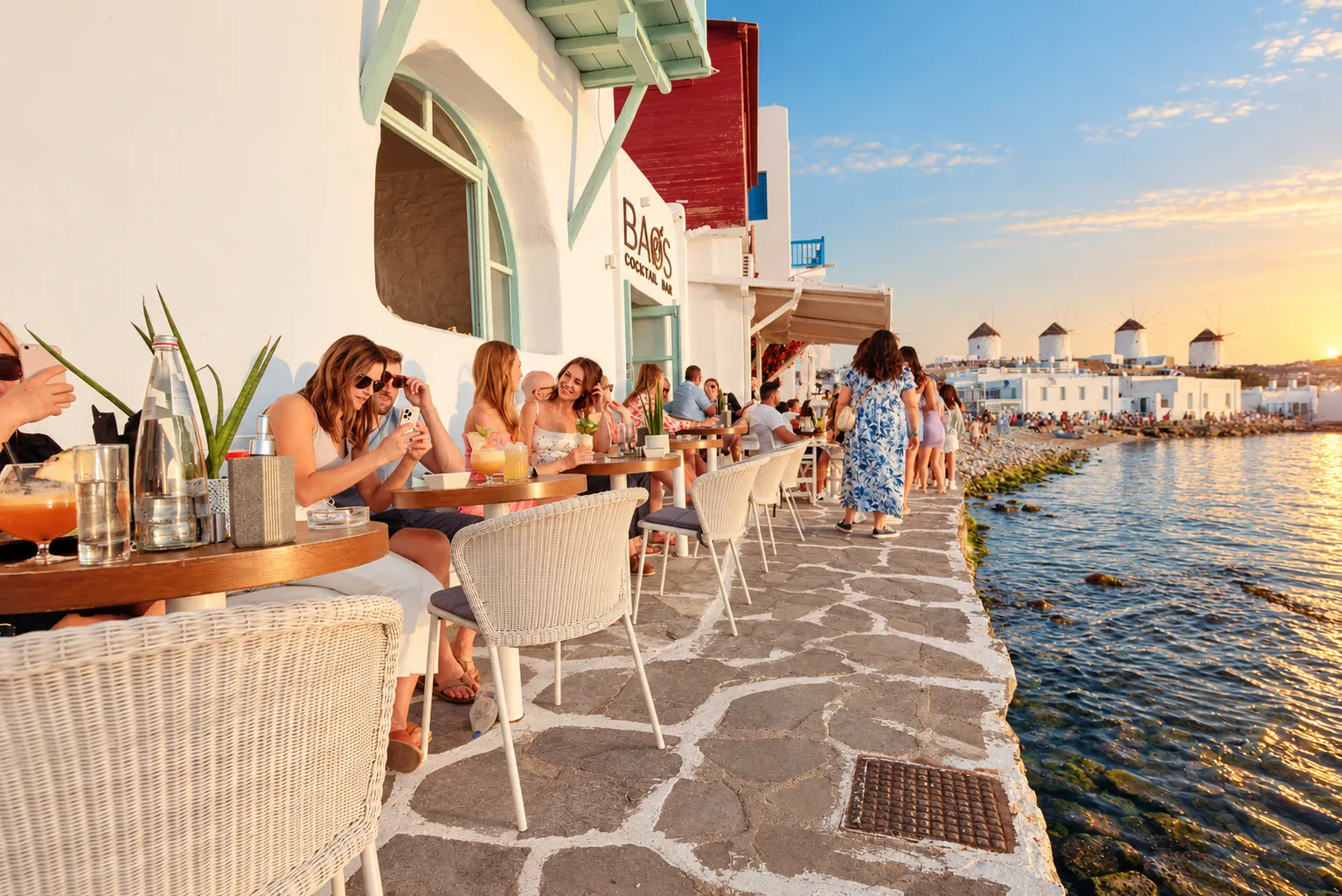 Gäste an den Tischen am Wasser im BAO'S in Klein-Venedig bei Sonnenuntergang, das Schild der Bar darüber und die Windmühlen auf der anderen Seite der Bucht