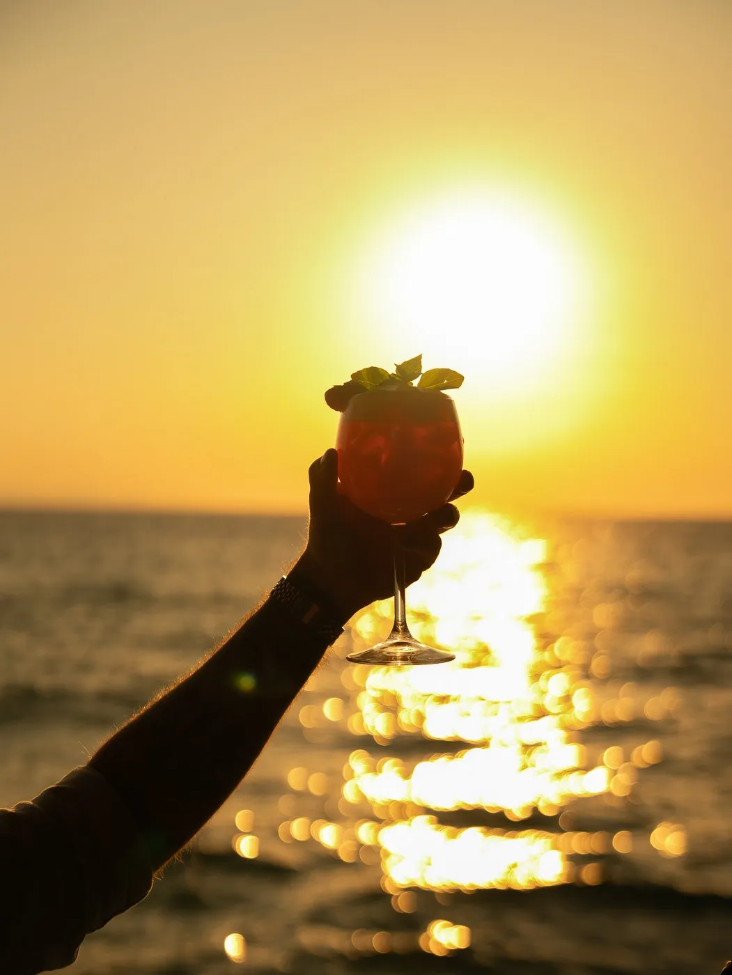 Ein erhobenes Glas vor der über dem Meer untergehenden Sonne, im BAO'S