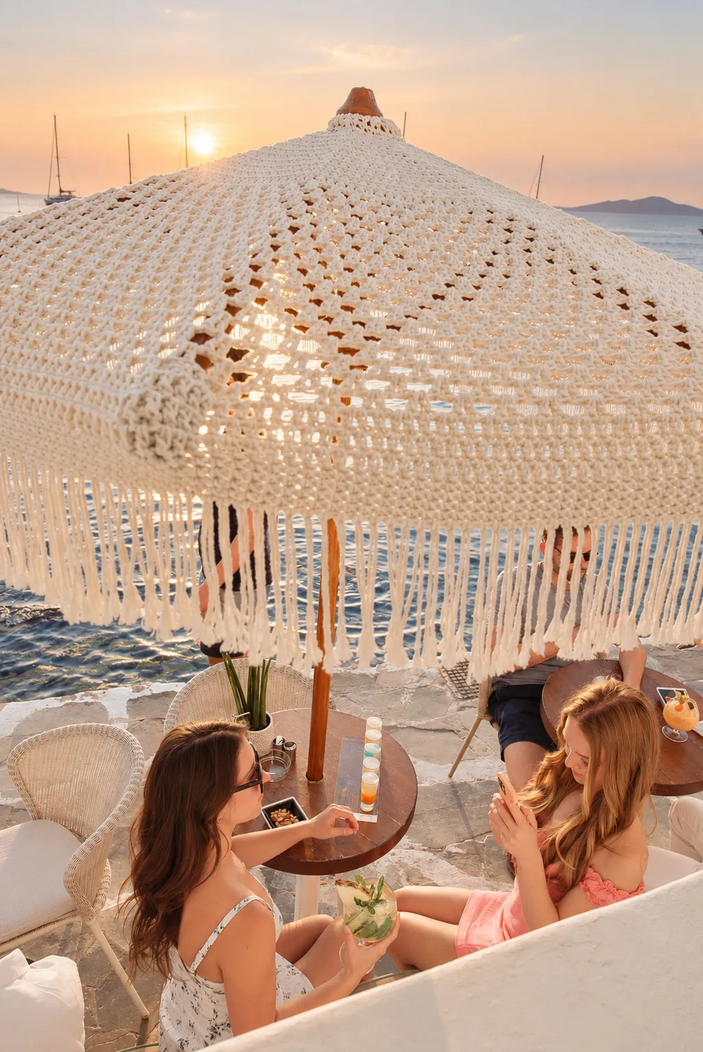 Two guests at a waterfront table at BAO'S under a macramé parasol, the sea and the sunset beyond